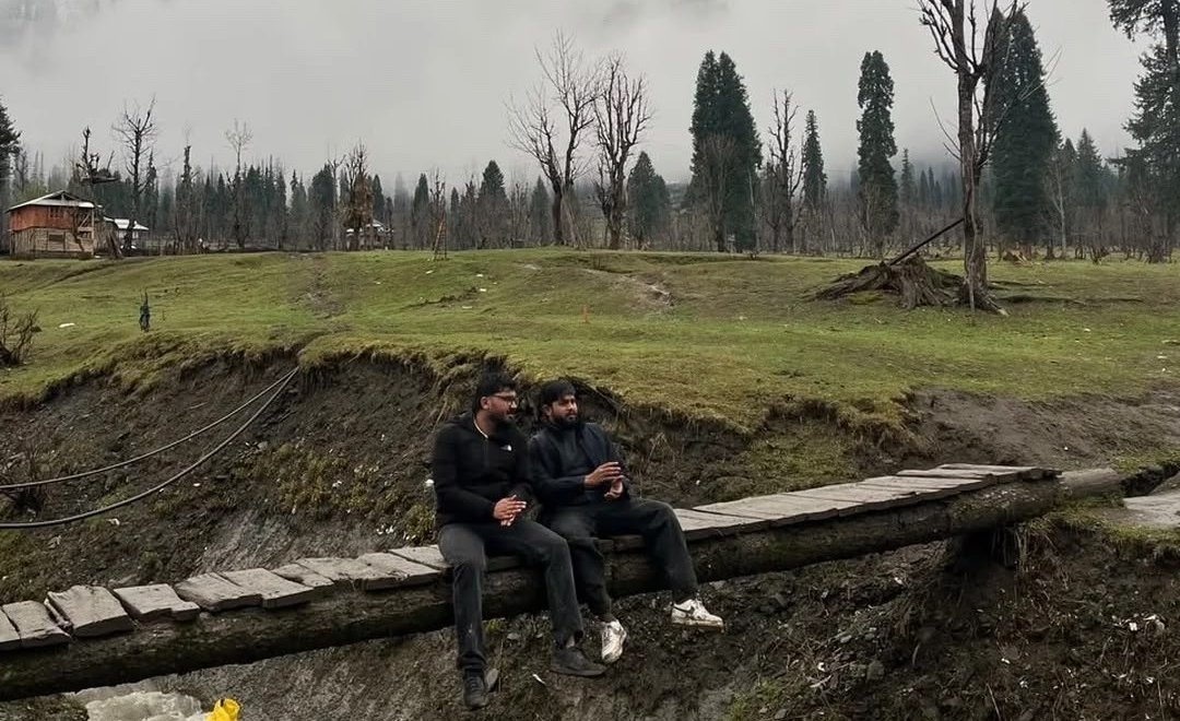 Two friends sitting on a wooden bridge over a stream in a misty Kashmir meadow